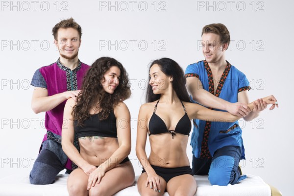 Theme massage and body care. Two brother Caucasian man in uniform make massage of hands and shoulders two beautiful sexy European women in underwear sitting on massage table isolated white background