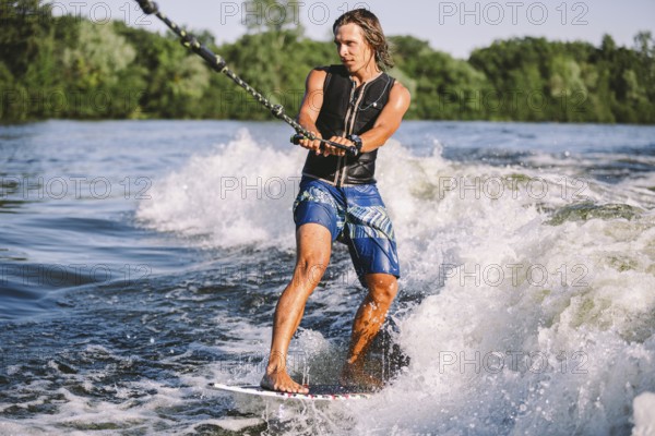 Beautiful long haired blond wakesurf in vest on board along waves of lake. Athletic male athlete wakes surfing in summer on river. Wakesurf water sport. Surfer on wave. Man surfing on the surfboard