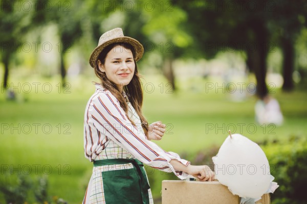 Photo theme small business cooking sweets. A young caucasian woman with an apron trader in the hat the owner of the outlet makes a candy raft, a fairy raft or a cotton candy in the summer park