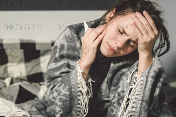 Subject disease and treatment. young Caucasian woman sits home bedroom bed wrapped in body blanket with high fever, fever holds strong headache behind head, uses drop medicine in nose for sinusitis