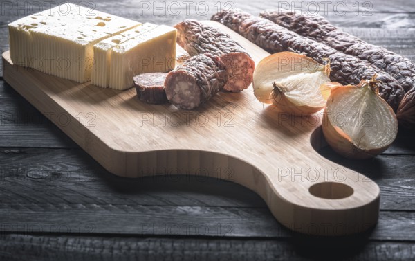 Tasty meal with homemade smoked sausages from beef and pork, fresh cheese and onion, displayed on a wooden cutting board, on a rustic black table