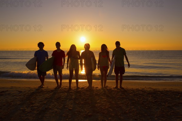 Surfers teen boys and girls group walking on beach at sunshine sunset backlight