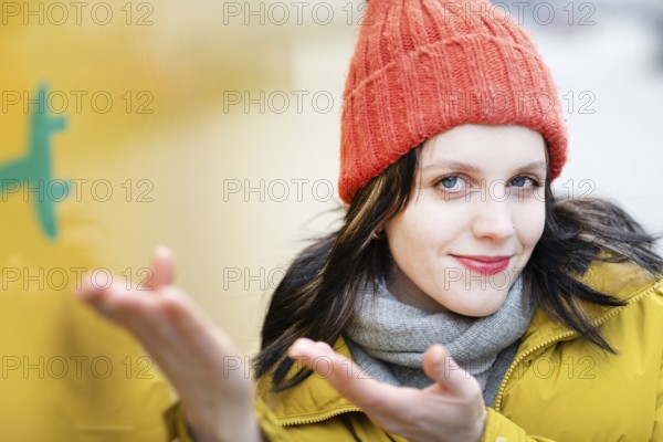 Woman with red hat and yellow jacket gestures to electric car for transport against urban background, environmentally friendly