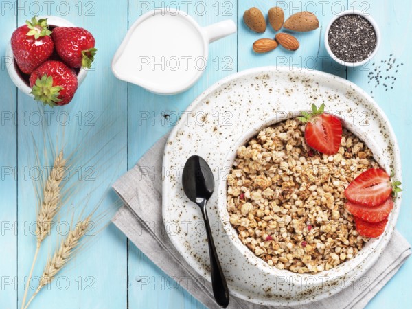 Top view of bowl with muesli, fresh strowberry, nuts, chia seeds and milk in milk jug on blue wooden background. Copy space