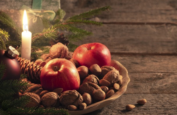 Christmas scenery with a wooden platter full of walnuts, hazelnuts and apples, green fir twigs and a gift box, on a vintage table, at candlelight