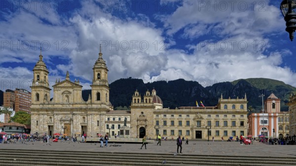 A historic cathedral and buildings surround a bustling plaza with people walking under a dramatic cloudy blue sky and mountain backdrop, Historic center, Bogotá, Colombia, South America