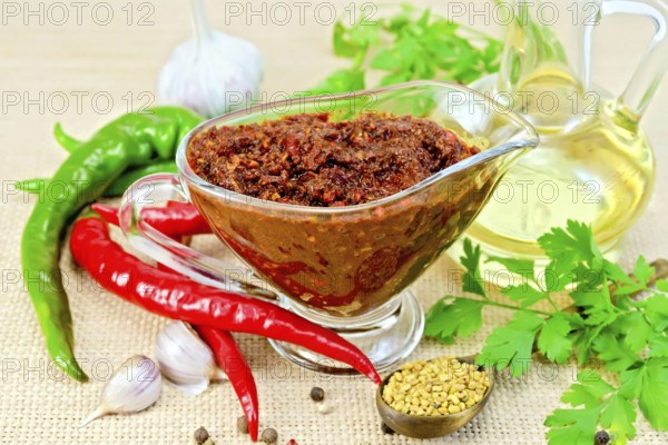 Tabasco adjika in a glass gravy boat, hot pepper, garlic, parsley, fenugreek, oil in the decanter on the background of sack cloth