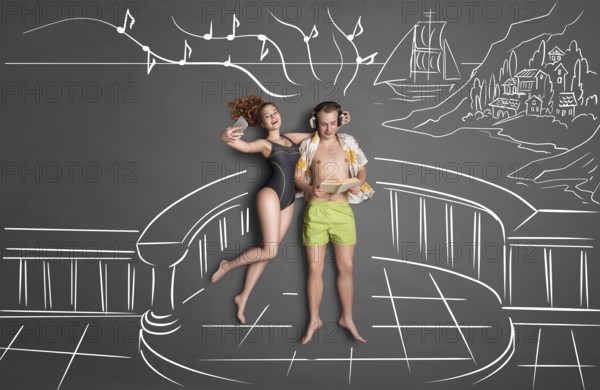 Love story concept of a romantic couple on the shore against chalk drawings background, view from above. Man wears headphones and reads a book, woman takes selfie with a smartphone