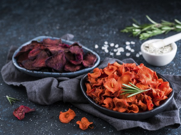 Baked beet and carrot slices with coarse sea salt scented rosemary. Vegan diet food idea and recipe. Healthy homemade beetroot and carrot chips in plate and ingredients on dark blue background. Copy space