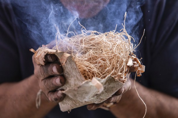 Closeup of man hand holding straw used as fire starter with smoke during demonstration of native traditional fire lighting in world and word festival
