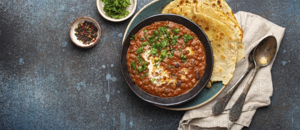 Traditional Indian Punjabi dish Dal makhani with lentils and beans in black bowl served with naan flat bread, fresh cilantro and two spoons on blue concrete rustic table top view. Space for text, food photography, food photography