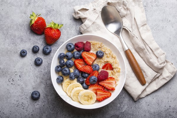 Oatmeal porridge with fruit and berries in bowl with spoon on white wooden background table top view, homemade healthy breakfast cereal with strawberry, banana, blueberry, raspberry, food photography