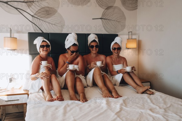 Four girls friends in white towels sitting on the bed having fun in hotel. Vacation Bloggers sunglasses drinking coffee together. Chat sweetly, tell stories and laugh. Friends met and spend their leisure time in hotel room. Concept of friendship