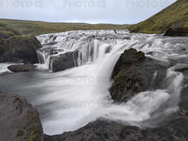 Water cascades over rocks into a flowing river in a serene natural setting. Iceland