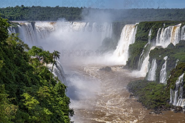 View to Devil's Throat and Iguazu Falls gorge with brown river, white cascading falls and lush green rainforest in sunshine