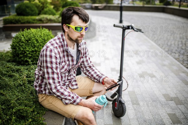 Man with e-scooter relaxing and using smartphone on bench in city. Modern male with electro scooter sitting on bench in park while chatting on his mobile phone. Student with eco transport hold phone