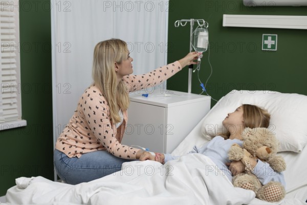 In a hospital, a caring mother supports her child with an IV, who holds a beloved teddy bear for comfort
