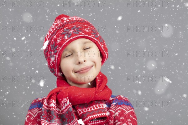 Dreamy child in warm clothing against a winter background. A boy with a red knitted hat and closed eyes. Fashionable boy in the studio in white snowflakes