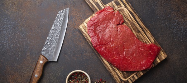 Raw uncooked top round beef steak on wooden cutting board with big kitchen knife and pepper on dark brown rustic stone background top view, cooking meat steak concept, food photography, food photography