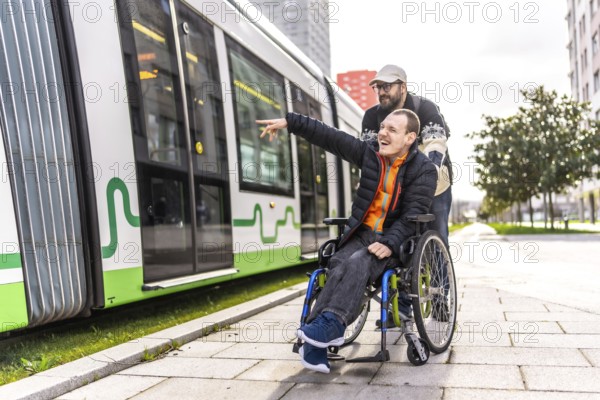 Disabled man in a wheelchair points at something while his friend stands behind him near a tram