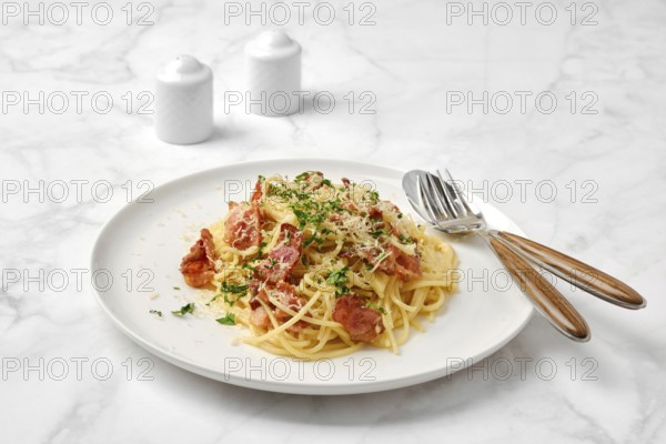 A delicious plate of spaghetti carbonara topped with crispy bacon, grated cheese, and fresh herbs on a marble table. Small salt and pepper shakers complete the casual dining setting