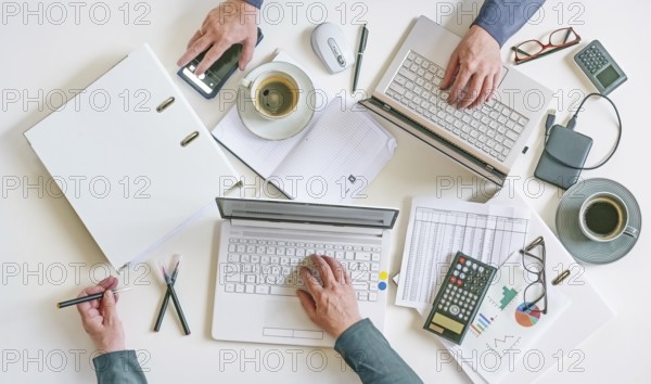Top view of a white office desk with hands of two people working on laptops with papers, calculator, and coffee, teamwork and accounting concept, high angle shot from above, copy space, selected focus