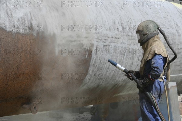 A man weating full safety gear sandblasts the steel casing of a meat drying machine for a rendering plant with ground glass