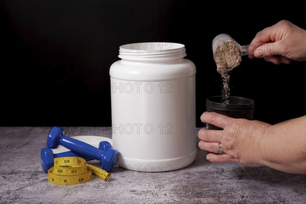 Woman pouring protein powder into a shaker with a spoon, next to a bottle of protein, dumbbells and a tape measure with a black background and copy space