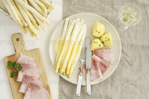 Traditional German dish with white asparagus, boiled ham, hollandaise sauce and potatoes, served on a light-coloured plate, accompanied by a glass of white wine