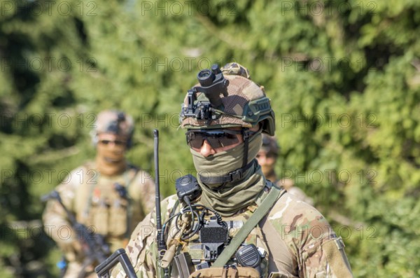 A soldier in camouflage uniform with tactical gear and sunglasses patrols a forest area