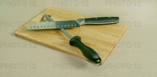 Santoku knife and honing steel on woodblock cutting board with light wood grain background