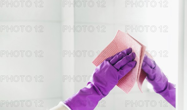 Cropped view of housekeeper woman hand in purple rubber gloves polishing mirror with pink cloth rag cleaner while cleaning bathroom at home. Housekeeping and cleaning service. Clean house, cleanliness