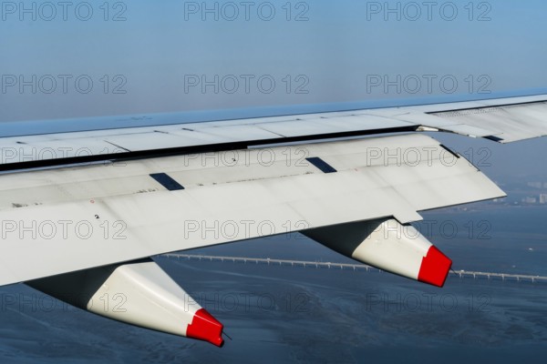 Aircraft wing with red tipped pillion in flight above ocean coastline with city buildings and bridge visible below