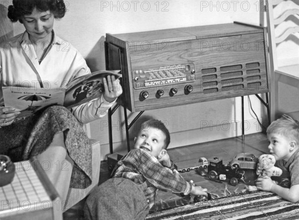 In the 1950s. A mother is seen sitting reading a magazine in her home while her sons are playing with toy cars on the floor. A typical 1950s radio-recordplayer with a wooden cabinet is seen. Sweden 1957