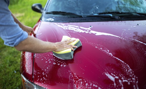 Hand using a sponge to wash the hood of a red car with soap and suds