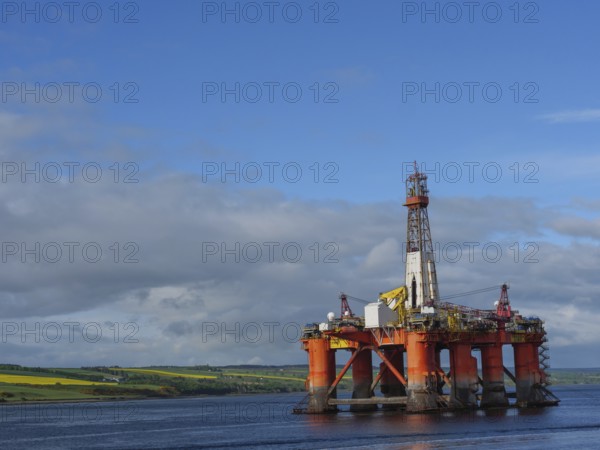 A single oil drilling platform sits in the calm sea with a slightly cloudy landscape behind it, drilling rig in red against a blue sky in the north sea, inverness, Scotland, United Kingdom, Europe