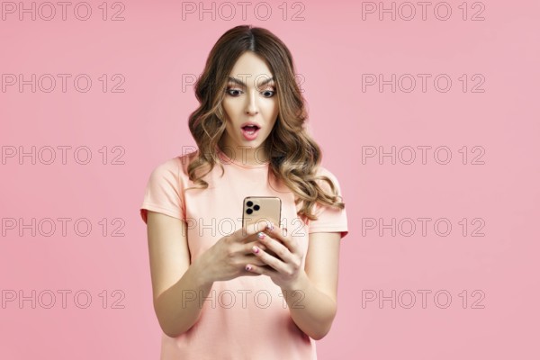 Portrait of a shocked pretty millennial hipster girl using mobile phone on pink background. surprised concept