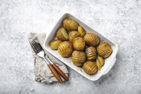 Hasselback potatoes with herbs in white ceramic casserole dish on white concrete table background from above. Delicious homemade accordion baked potatoes for dinner, food photography, food photography