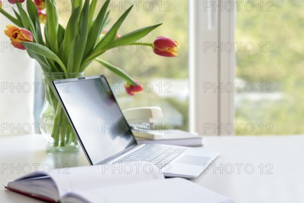 Laptop with organizer and a flower bouquet from tulips on a white desk by the window, business in the home office, copy space, selected focus, narrow depth of field