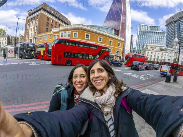 Two happy female tourists taking a selfie with red double decker buses and modern skyscrapers in the background, enjoying their vacation in london