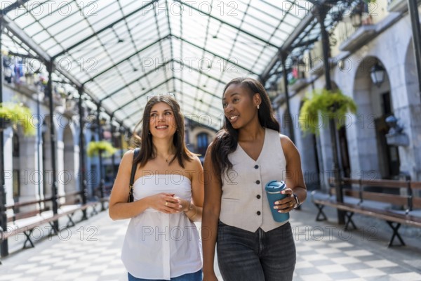 Two young businesswomen are walking and talking in a covered gallery, enjoying a break and holding reusable coffee cups