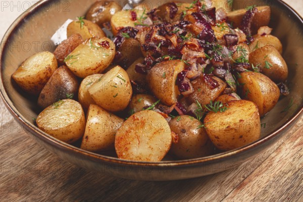 A bowl filled with golden roasted potatoes seasoned with spices and garnished with caramelized onions and fresh herbs sits on a wooden table, showcasing a flavorful dish prepared for a meal