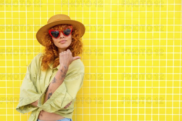 Fashionable influencer posing confidently against a vibrant yellow tiled wall, pointing with her thumb while showcasing her stylish look