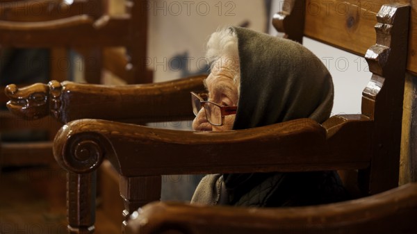 An elderly woman with a hood, sitting in a chair, deep in thought, Easter 2025, Church of the Dormition of the All-Holy God Bear, Kimisis tis Theotokou, Asklipio, Rhodes, Dodecanese, Greek Islands, Greece, Europe