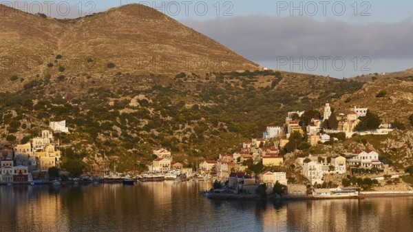 View of a settlement on a hill with warm light, calm water in the foreground, Symi Town, Symi, Dodecanese, Greek Islands, Greece, Europe