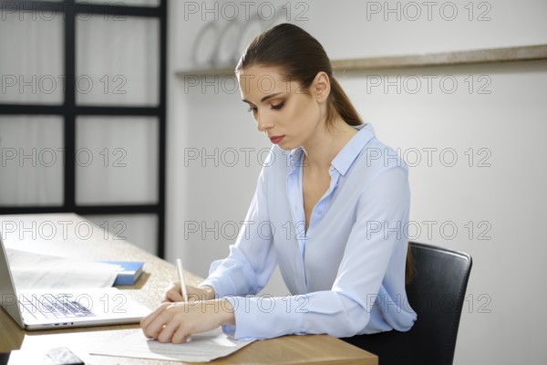 A business woman sits at a wooden desk, intently writing on a piece of paper while using her laptop. The bright office environment highlights her concentration and dedication to her tasks
