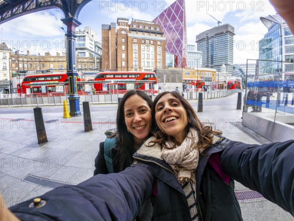 Two happy tourists taking a selfie in the city of london, with red buses and modern buildings in the background