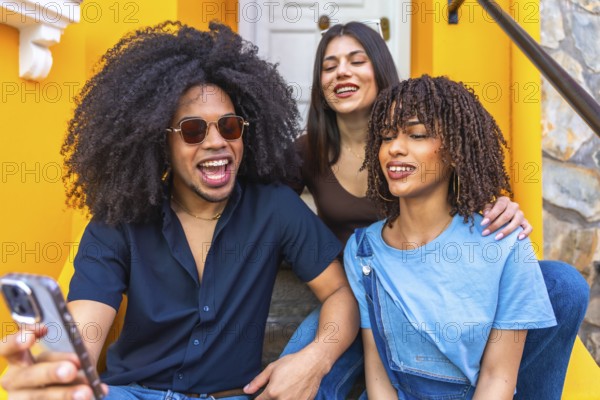 Three happy young friends are taking a selfie with a smartphone while sitting on the stairs of a colorful building