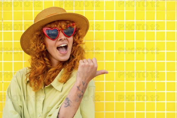 Excited young woman with red hair, heart shaped sunglasses, and straw hat pointing her thumb to the side while standing against a vibrant yellow tiled wall