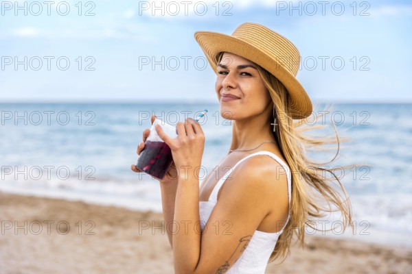 Young woman in a straw hat enjoying a refreshing drink while relaxing on the beach, soaking up the sun and savoring the summer vibes